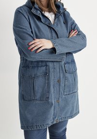 Džínová bunda ve středně modré barvě s kapucí, předním zapínáním na cvoky, stahovacími šňůrkami v pase a dvěma velkými předními kapsami. Obsahuje viditelné švy.