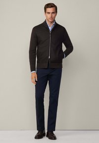 Veste marron foncé avec fermeture éclair, texture côtelée, col montant et coupe ajustée, associée à un pantalon bleu marine et des chaussures marron.