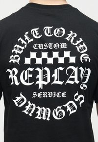 Άτομο που φοράει μαύρο πουκάμισο με λευκό γοτθικό κείμενο «Balt toride», «Custom», «Replay» και «Service» γύρω από ένα μοτίβο σκακιέρας.