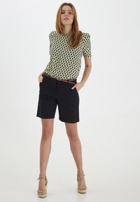 T-shirt à manches courtes de couleur vert clair avec un motif géométrique noir ; associé à un short bleu marine et des talons compensés beiges. Détails notables : une silhouette ajustée.