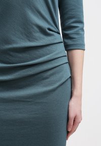 Robe bleu sarcelle en tissu doux et texturé, avec des manches trois-quarts et des détails froncés à la taille. Gros plan sur une main posée sur le tissu.