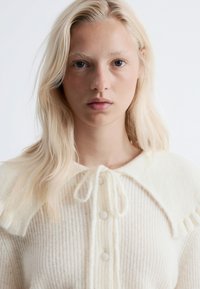 Helle beige Strickjacke mit rundem Kragen, gewellten Rändern und Knöpfen vorne. Weiche Textur mit einem dezenten Streifenmuster.