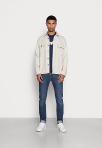 Vaaleanharmaa nappikauluspaita mustilla nappuloilla, kahdella rintataskulla, päällä tummansinisellä t-paidalla, yhdistettynä tummansinihin farkkuihin ja valkoisiin lenkkareihin.