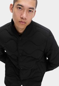 Svart quiltad jacka med vågmönster, tryckknappar, ribbad krage och mudd. Tyget verkar vara lättviktigt med en jämn yta.