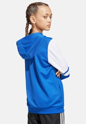 Jeune femme aux cheveux tressés tournant la tête, portant un sweat à capuche bleu avec des manches blanches et un pantalon noir à rayures blanches.