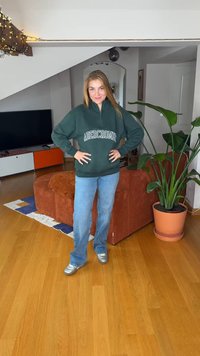 Groene sweatvest met ritssluiting en witte tekst, blauwe wijde spijkerbroek en zilveren schoenen. Binnenomgeving met een plant en een bruine bank op de achtergrond.