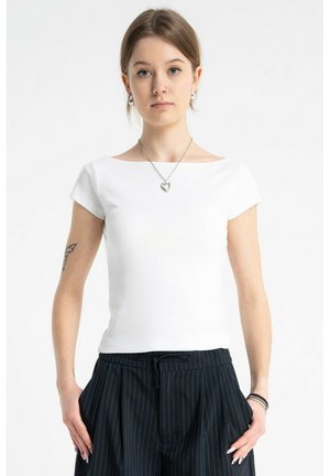 Młoda kobieta o krótkich włosach, ubrana w białą dopasowaną bluzkę, czarne spodnie w prążki, z zawieszką w kształcie serca i widocznym tatuażem na ramieniu.