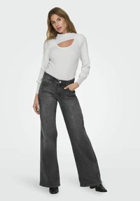 Weißer Ripp-Pullover mit Ausschnitt-Kragen, kombiniert mit hoch taillierten, weiten, dunkelgrauen Jeans. Das Modell steht vor einem neutralen Hintergrund.