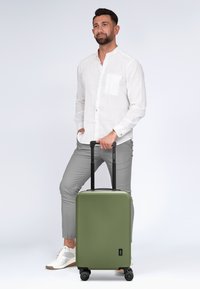 Grünes Hartschalentrolley mit glatter, matter Oberfläche, schwarzem Teleskopgriff und vier Rädern für Mobilität. Minimales Design, kompakte Form.