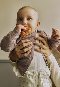 Bērniņš, tērpies adītā lavandas krāsas kardiganā, tur cepumu, ko ieskauj rokas. Bērniņš valkā baltu ribotu krekliņu un bikses ar ziedu rakstu.