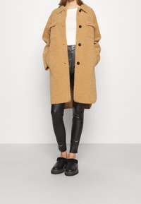 Tan, textured oversized coat with two front pockets and black buttons, paired with shiny black leggings and black fur-accented shoes.