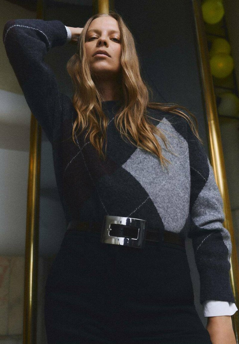 Maglione scuro grigio a motivo argyle realizzato in tessuto testurizzato, abbinato a pantaloni neri a vita alta e una cintura metallica con fibbia larga.