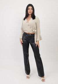 Top enveloppant beige avec des accents de boutons, associé à un pantalon en simili cuir noir taille haute. Modèle portant des sandales à talons transparents.