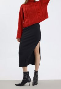 Pull à col roulé rouge en maille torsadée, court, associé à une jupe midi noire avec une fente haute sur la cuisse, et des bottines noires à talons pointus.