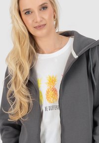 Felpa grigia con zip abbinata a una maglietta bianca con grafiche colorate di ananas. Tessuto morbido, vestibilità rilassata e dettagli del cappuccio.