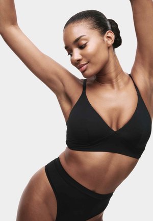 Kobieta z włosami związanymi w kok, ubrana w czarny biustonosz i majtki z wysokim stanem, podnosi ramiona i spogląda w dół z lekkim uśmiechem.