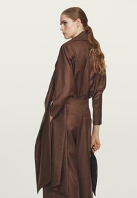 Brązowa, dopasowana sukienka koszulowa z długim rękawem, z paskiem w talii, bocznymi rozcięciami i gładkiego materiału. Uzupełniona ciemną torebką kopertową.