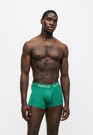 Muskelbærer mand iført grønne Nike boxershorts, stående med front mod kameraet med et neutralt ansigtsudtryk mod en ensartet lys baggrund.