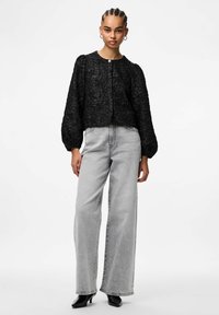 Sort teksturert kort jakke med puffede ermer, kombinert med lysegrå vidbukser og svarte sko med hæl.