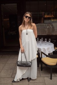 Biała, bezramiączkowa, asymetryczna sukienka z płynącym materiałem; kobieta trzyma czarną torebkę z plecionymi uchwytami, nosi okulary przeciwsłoneczne i szpilki.