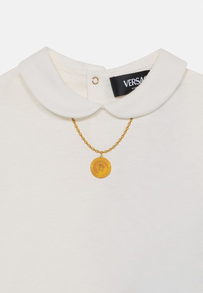Balta apykaklė marškiniai su apvalia iškirpte, puošti auksine grandine ir apvaliu pakabuku. Etiketė skelbia "VERSACE."