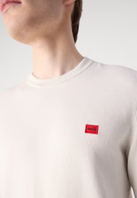 T-shirt en coton beige avec un col rond et des manches courtes, arborant un petit logo rectangulaire rouge brodé sur la poitrine.