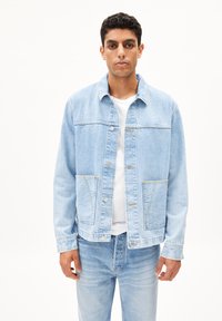 Lichtblauwe denimjack, relaxte pasvorm, twee voorzakken, zilveren knopen met een metallic uitstraling, contrasterende stiksels, gecombineerd met een wit T-shirt en bijpassende jeans.