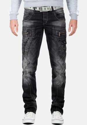 Mann trägt ausgewaschene schwarze Cargohosen mit aufgenähten Details, Reißverschlusstaschen, weißem Gürtel, grauem Sweatshirt und weißen Sneakers, steht aufrecht.