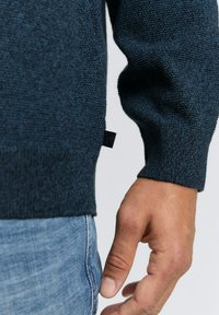 Dunkelblauer Strickpullover mit strukturierter Oberfläche und gerippten Bündchen, mit einem kleinen Etikett an der Seite. Getragen mit hellblauen Jeans.