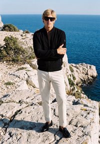Homme blond portant une veste noire, un pantalon blanc et des lunettes de soleil, debout les bras croisés sur une falaise rocheuse au bord de la mer sous un ciel dégagé.