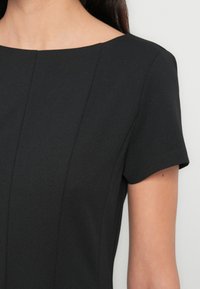 Gros plan d'une personne portant une robe noire à manches courtes avec un col carré et des détails de couture verticale à l'avant.