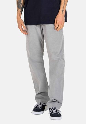 Person wearing light gray loose pants and black sneakers with white laces, standing with hands at sides showing tattoos on arms.