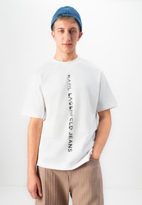 Jaunas vyras vilki baltą marškinėlį su vertikalia „KARL LAGERFELD JEANS“ užrašu, smėlio spalvos kelnes ir mėlyną džinsinį kepuraitę atvirkščiai.