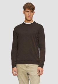 Brauner Rundhalsausschnitt-Pullover mit langen Ärmeln und gerippten Bündchen. Mit einem subtilen Logo auf der Brust. Über einem weißen Hemd getragen.