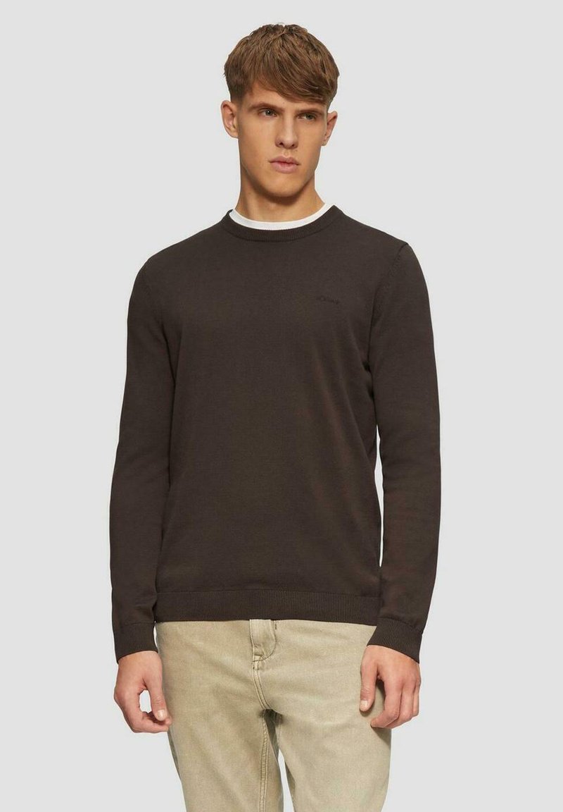 Brauner Rundhalsausschnitt-Pullover mit langen Ärmeln und gerippten Bündchen. Mit einem subtilen Logo auf der Brust. Über einem weißen Hemd getragen.