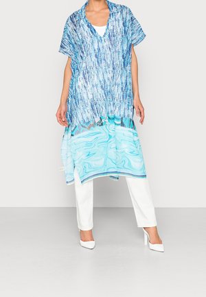 Personne portant une tunique bleue à motifs sur un pantalon blanc et des talons blancs, debout devant un fond clair uni.