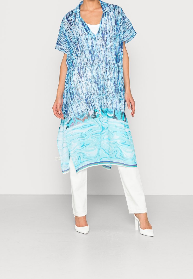 Personne portant une tunique bleue à motifs sur un pantalon blanc et des talons blancs, debout devant un fond clair uni.