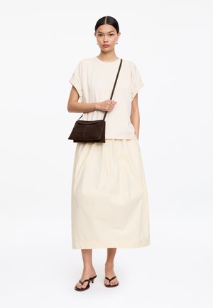 Nainen, jolla on tummat hiukset, kermaisen värinen lyhythihainen paita ja siihen sopiva pitkä hame. Hän kantaa tummanruskeaa olkalaukkua ja käyttää mustia korkokenkiä.