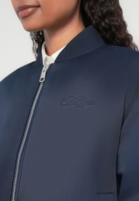 Navy bomber jacket with a zip closure, ribbed collar, and tonal embroidered logo. Smooth, lightweight fabric with a sleek design.