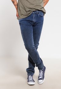 Vaqueros azules de mezclilla con un ajuste ajustado, que presentan una cintura de tiro medio, piernas rectas y un sutil desgaste. Combinados con zapatillas blancas.