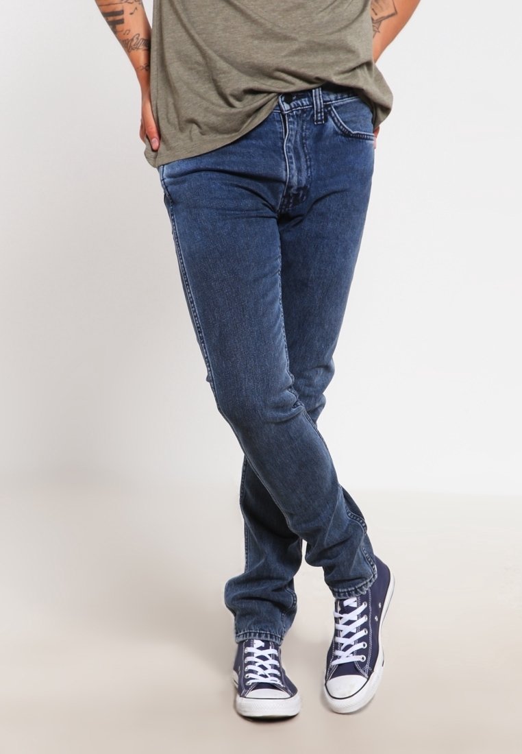 Vaqueros azules de mezclilla con un ajuste ajustado, que presentan una cintura de tiro medio, piernas rectas y un sutil desgaste. Combinados con zapatillas blancas.