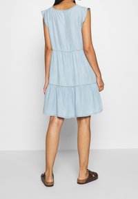 Vaaleansininen, lyhythihainen mekko, jossa on röyhelöreunat, kerroksellinen muotoilu ja rento istuvuus, valmistettu denimiä muistuttavasta kankaasta. Ruskeat sandaalit näkyvät alla.