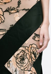 Fekete szövet áttetsző réteggel, amely barack színű virágmintázatot és fekete kontúrokat tartalmaz. A varrásnál elhelyezett kéz éles, szögletes anyagrészeket mutat.