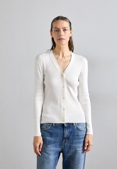 Cardigan blanc à côtes avec un col en V et une fermeture à boutons, doté de manches longues et d'une coupe ajustée, associé à un jean en denim bleu.