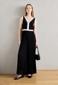 Combinaison noire plissée avec un accent blanc à la taille et un décolleté en V, assortie avec des ballerines noires et un petit sac à main noir. Texture de tissu lisse.