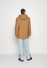 Brauner Wollmantel mit Kapuze, entspannter Schnitt und knöpfbaren Ärmeldetails. Getragen über hellblauen Jeans und schwarzen Plateau-Stiefeln.