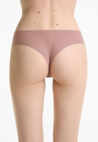 Lichtgetinte persoon in naadloze mauve stringondergoed, met zichtbare onderrug, billen en bovenbenen tegen een witte achtergrond.