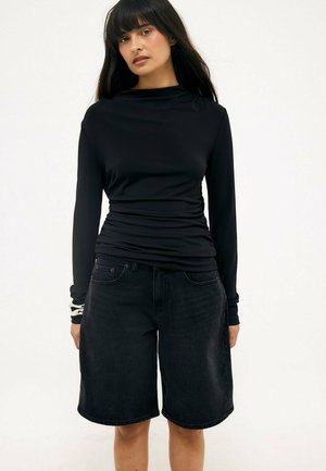 Kvinde med langt mørkt hår iført en tætsiddende sort bluse med lange ærmer og løse sorte denim knælange shorts på hvid baggrund.