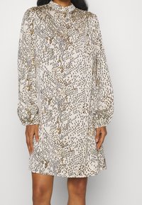 Robe à manches longues, longueur genou en tissu léger avec une base beige et des motifs abstraits noirs et dorés, présentant un col droit et des manches froncées.