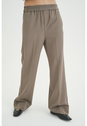 Løse taupebukser med elastik i taljen båret med sorte spidse flade sko på en stående person mod en hvid baggrund.